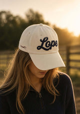 vit keps med mörkblå lope text i vintage stil i filt på fronten. broderad äst i brint på sidan och texten "saddle up" bak i mörkblå brodyr. för ryttare
