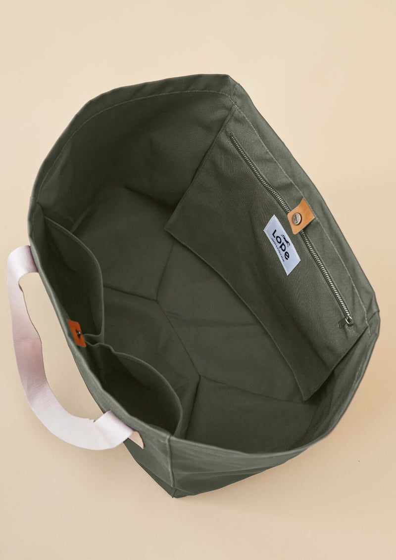 Lope Tote Bag Gröngrå stallväska. Tyg: 900D polyester i grågrön färg. Axelband: Vävda band i crèmevitt med nedsänkt text LOPE SPORT. Sömmar: SPI (stitches per inch): 8–9. Foder: Dubbelt tyg (samma polyester även som insida). Tråd: Nylon, med hög slitstyrka. Logo-lapp: Cognacsfärgat PU-läder med svart nedsänkt präglad logotyp. Stängningsrem: Cognacsfärgat PU-läder med tryckknapp i antik mässing präglad med logotyp. Uppifrån foto