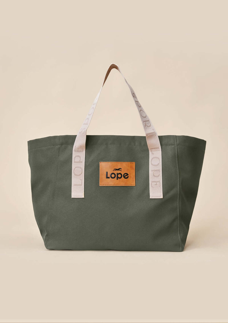 Lope Tote Bag Gröngrå stallväska. Tyg: 900D polyester i grågrön färg. Axelband: Vävda band i crèmevitt med nedsänkt text LOPE SPORT. Sömmar: SPI (stitches per inch): 8–9. Foder: Dubbelt tyg (samma polyester även som insida). Tråd: Nylon, med hög slitstyrka. Logo-lapp: Cognacsfärgat PU-läder med svart nedsänkt präglad logotyp. Stängningsrem: Cognacsfärgat PU-läder med tryckknapp i antik mässing präglad med logotyp