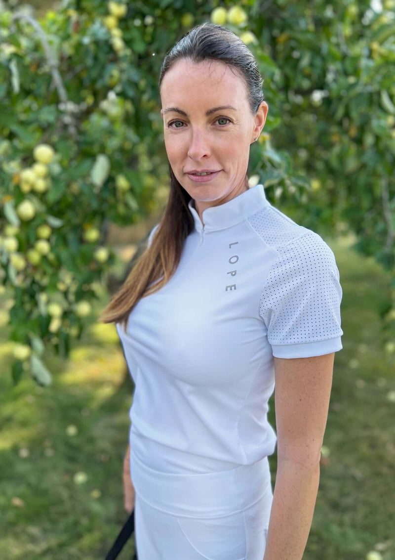 Alt-text: GRACE TOP är en vit, kortärmad tävlingstopp för ryttare, designad för optimal rörelsefrihet och en sportig, fräsch look. Den har en vit krage med dold dragkedja, korta ärmar för varmare dagar, och mesh i rygg och ärmar för ökad ventilation och komfort. Sportiga detaljer inkluderar en LOPE-logga i 3D-silikon framtill och en signaturhäst i nacken. Funktioner som snabbtorkande material och fyrvägsstretch gör den till ett utmärkt val för den moderna tävlingsryttaren.