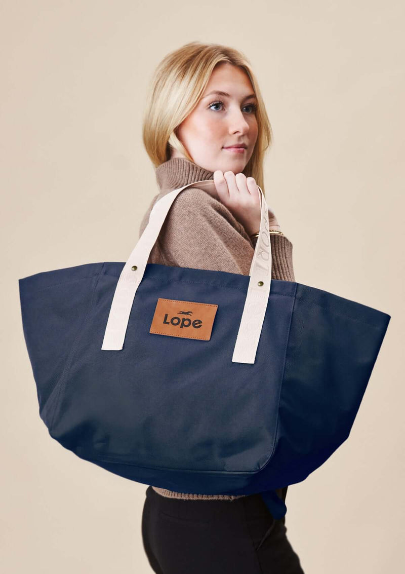 Lope Tote  midnattsblå– din nya favoritväska till stallet, jobbet och träningen. Tyg: 900D polyester i blåsvart färg. Axelband: Vävda band i crèmevitt med nedsänkt text LOPE SPORT med en nit i antik mässing Foder: Dubbelt tyg (samma polyester även som insida). Tråd: Nylon, med hög slitstyrka. Logo-lapp: Cognacsfärgat PU-läder med svart nedsänkt präglad logotyp.