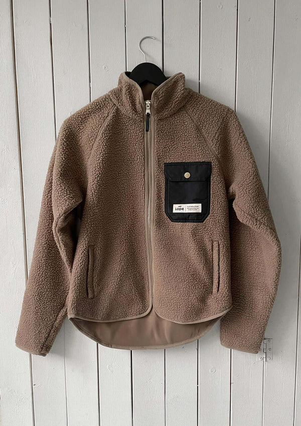 En beige fleecejacka med windstopper-funktion, hel dragkedja framtill och en svart bröstficka med präglad guld knapp och etikett. Jackan har hög krage, två sidofickor med dragkedja och en längre rundad nederkant baktill