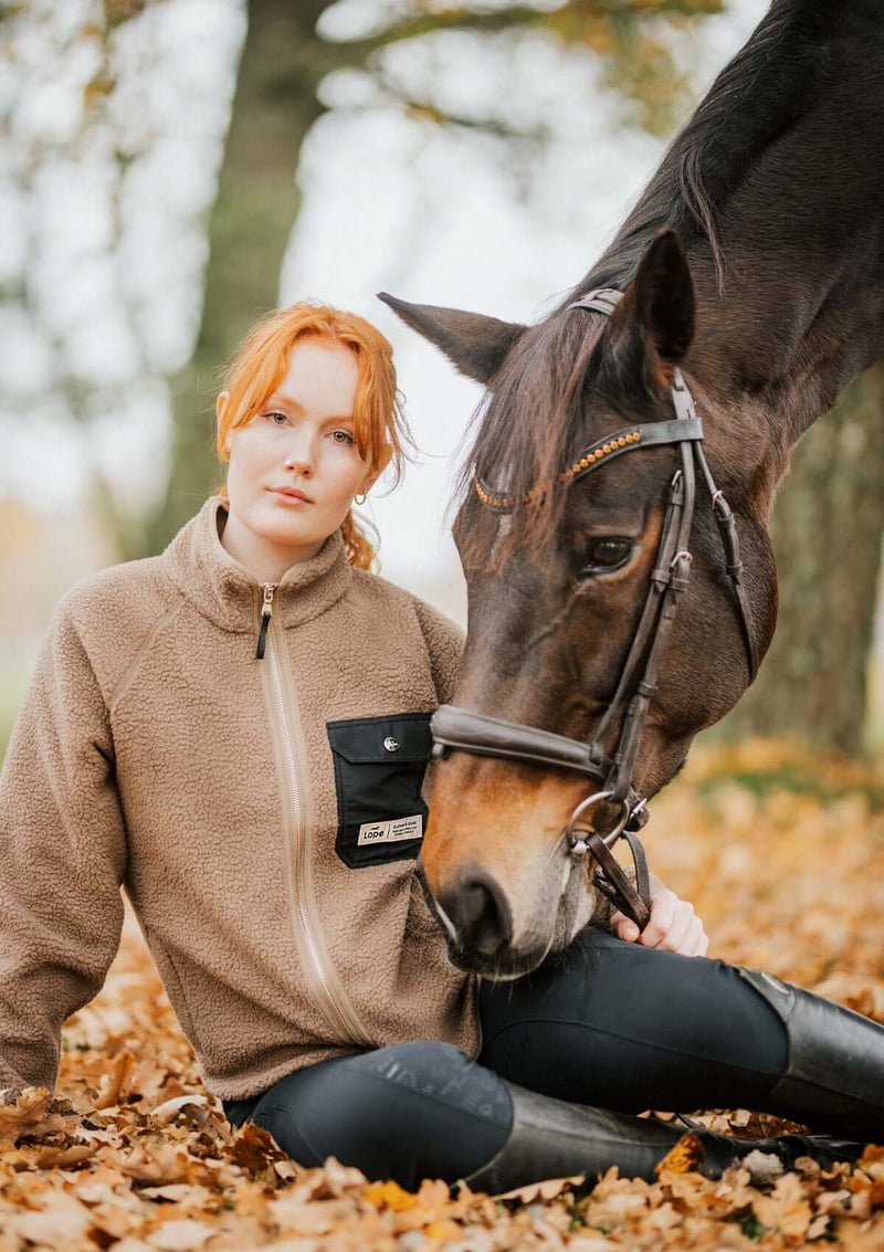 Vintät stalljacka i teddy fleece. Varm beige pilé fleece med raglanarm, tumhål, dolda sido fickor med blixtlås och en svart dekorativ ficka på bröstet och detaljer i ljust guld. rundad avslutning på jackan ger en kvinnlig passform.