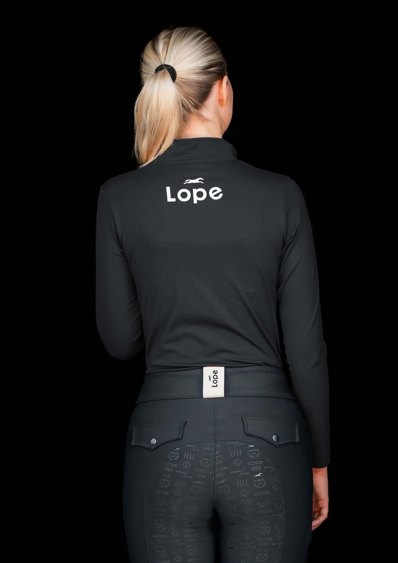 Kvinna i ridkläder: Svart långärmad quarter zip top i funktionsmaterial med 'lope' logotyp på ryggen och 'lope sport' logotyp på armen